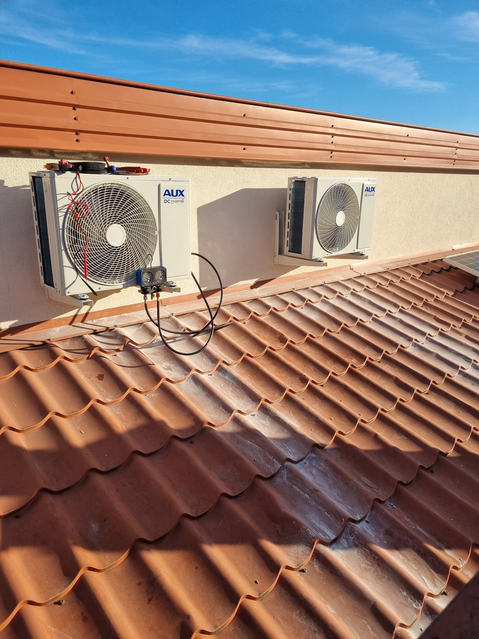 Dwa zamontowane na elewacji budynku klimatyzatory AUX DC Inverter, widoczne narzędzia pomiarowe podłączone do jednego z urządzeń, dachówka ceramiczna w tle.
