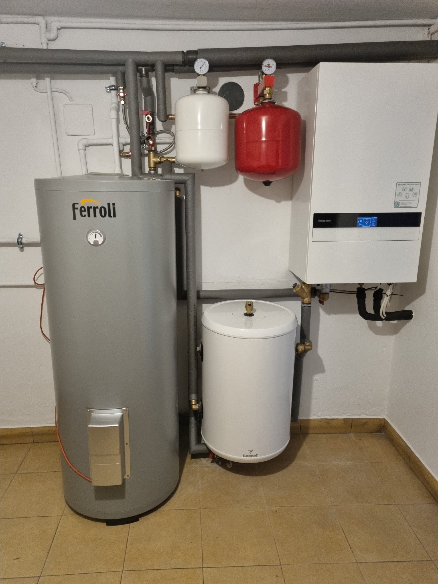 Instalacja kotła gazowego Panasonic Aquarea Smart Cloud obok szarego zbiornika Ferroli i białego zbiornika Galmet, z czerwonym i białym naczyniem przeponowym, rury instalacyjne na ścianie i brązowe...