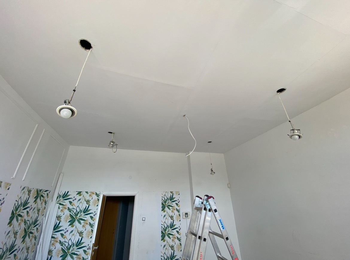 Wnętrze w trakcie montażu oświetlenia sufitowego, widoczna drabina aluminiowa, fragment tapety w kwiaty oraz wiszące przewody i oprawy żarówek.