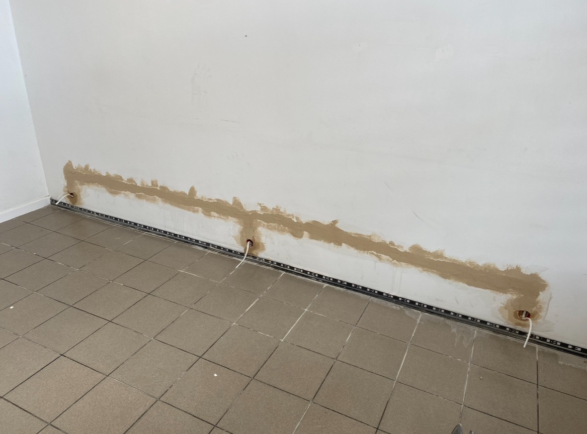 Ściana po bruzdowaniu pod przewody elektryczne, widoczne otwory i zamaskowane kable z użyciem gipsu szpachlowego, listwa przypodłogowa z ledami, płytki podłogowe.