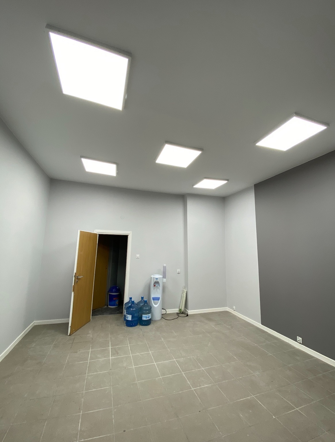 Wnętrze biurowe po montażu oświetlenia LED: widoczne kwadratowe panele LED w suficie, szare ściany, dystrybutor wody z butlami, otwarte drzwi do pomieszczenia gospodarczego.