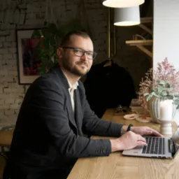 Mężczyzna w okularach i marynarce pracuje na laptopie przy drewnianym biurku w kawiarni z ceglaną ścianą w tle.