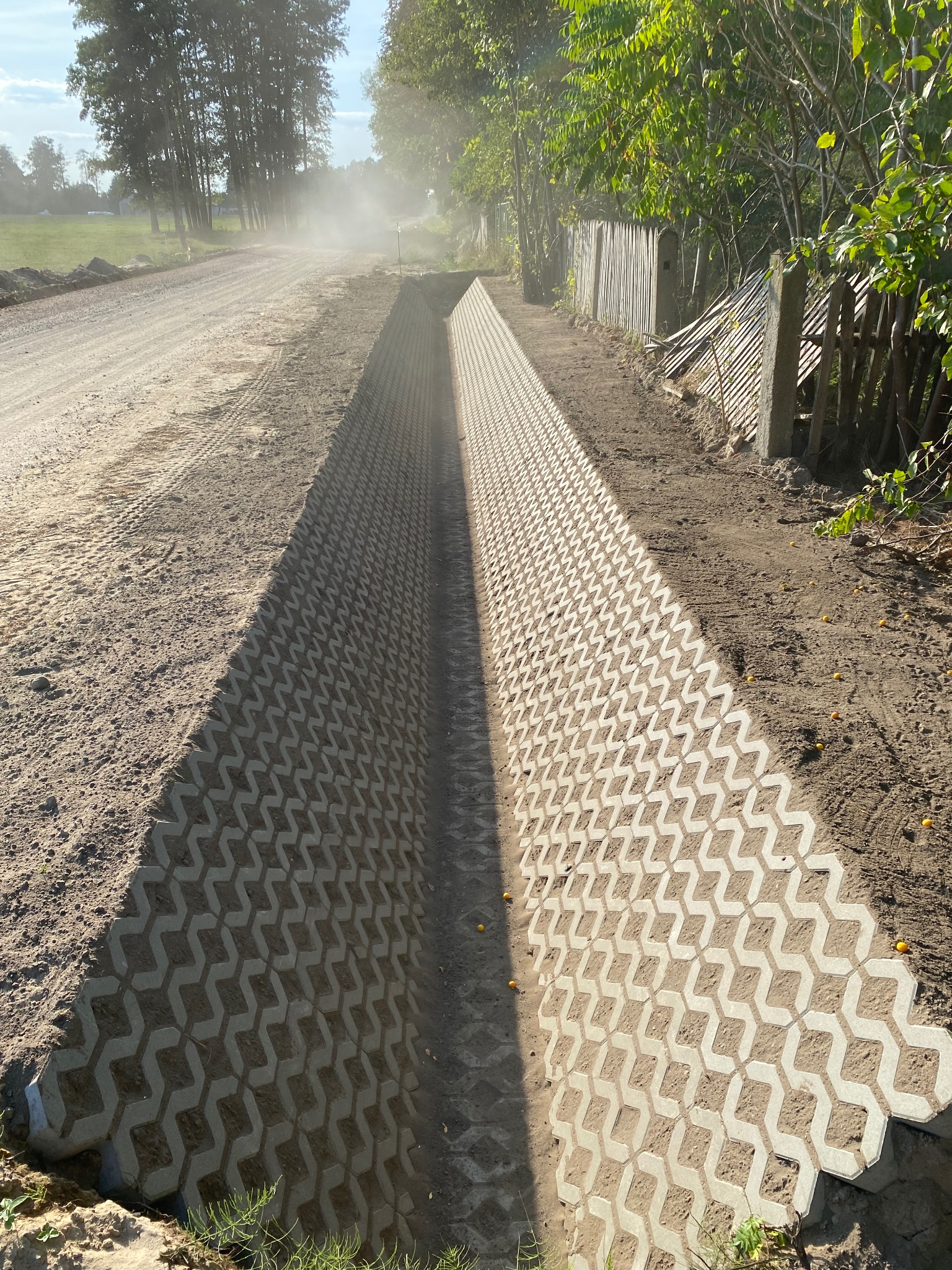 Koryto odwadniające z ażurowych płyt betonowych, widok z góry, Mińsk Mazowiecki. Ułożone wzdłuż drogi gruntowej, w tle drzewa i drewniany płot.