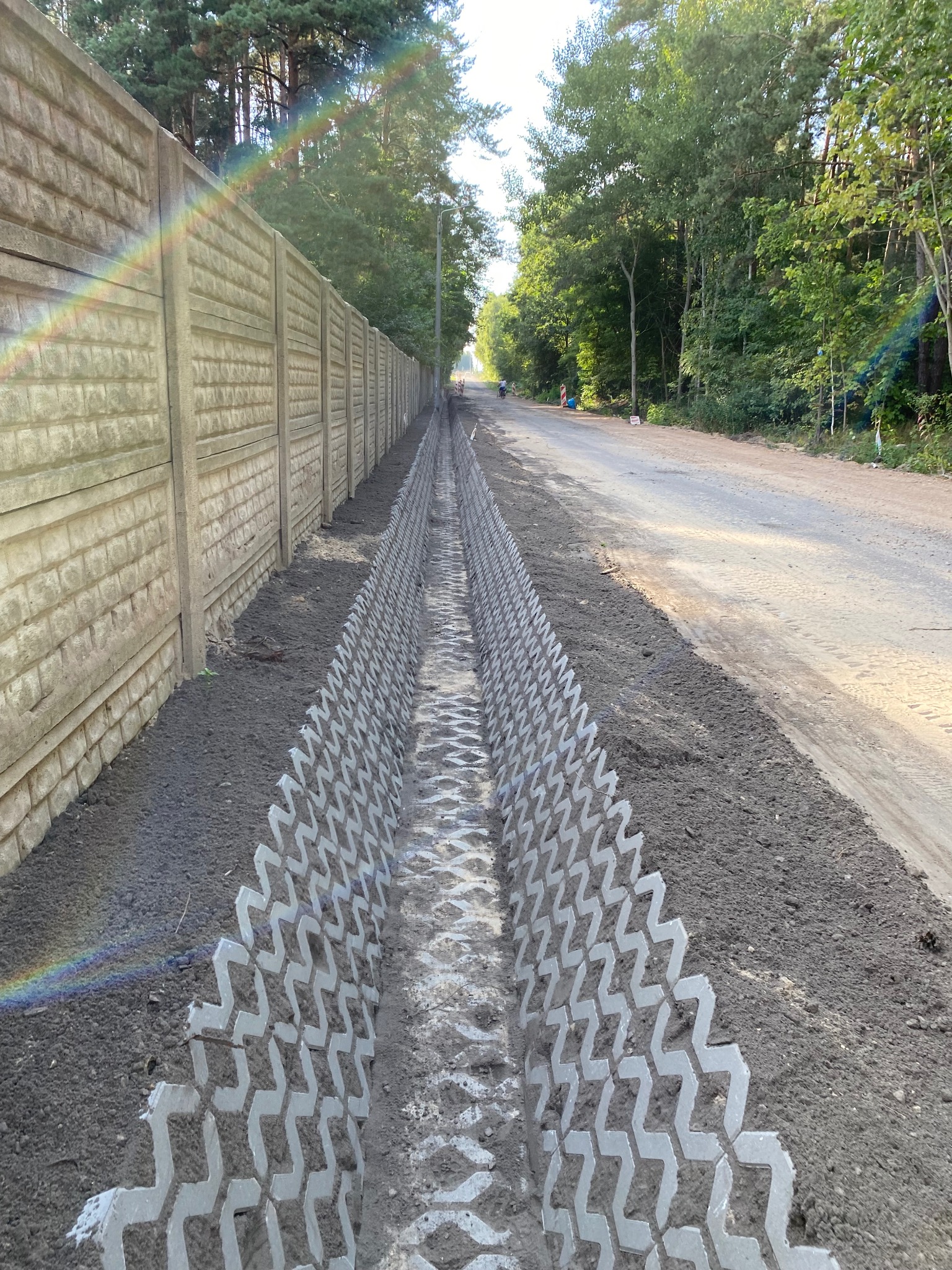 Długi rów wyłożony ażurową kostką betonową, biegnący wzdłuż drogi i betonowego ogrodzenia w otoczeniu drzew. Widoczny fragment tęczy.