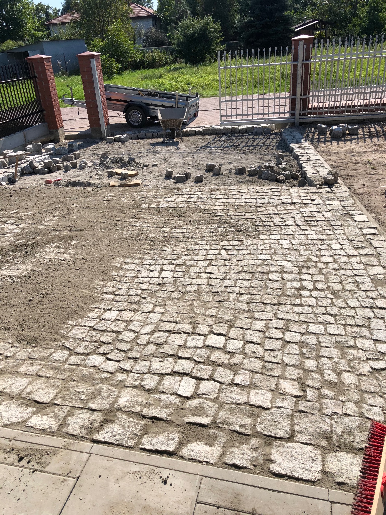 Fragment układanej nawierzchni z kostki granitowej na posesji. Widoczny fragment ułożonej kostki, przygotowany teren i materiały budowlane. W tle brama wjazdowa.