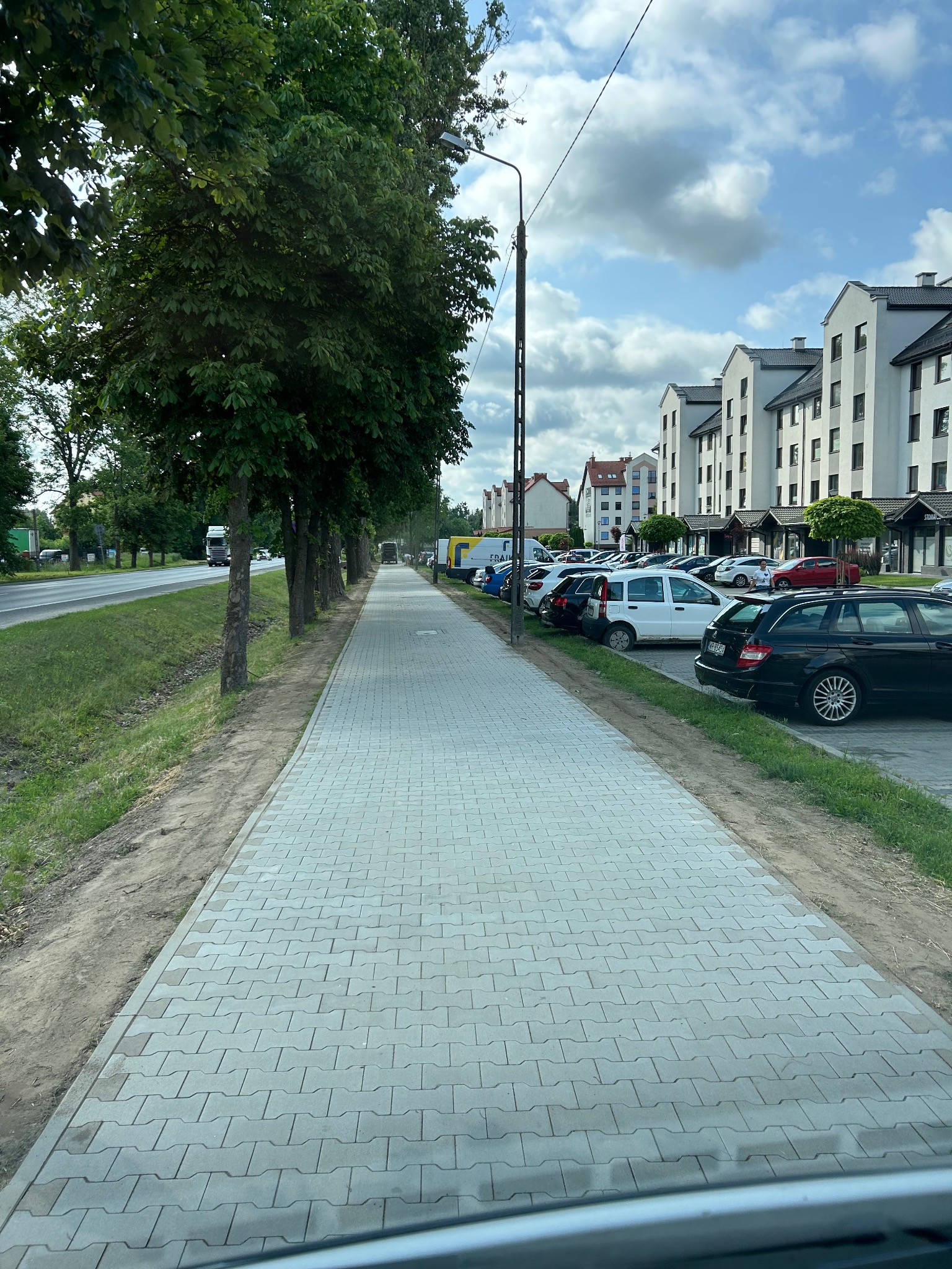 Długa, szara ścieżka z kostki brukowej, ciągnąca się wzdłuż ulicy z drzewami i zaparkowanymi samochodami. Tło stanowią budynki mieszkalne i błękitne niebo z chmurami.