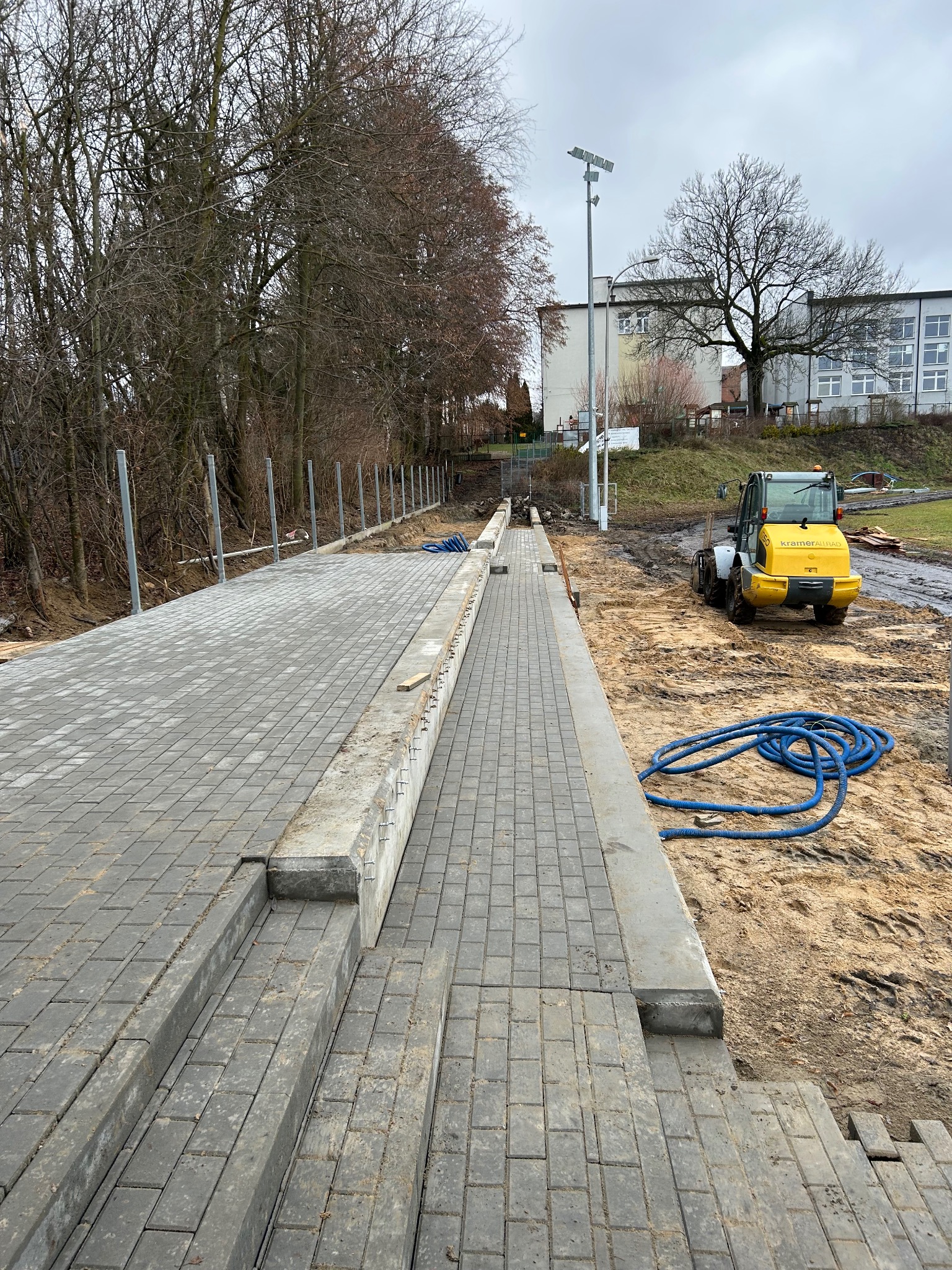 Fragment układanej szarej kostki brukowej z obrzeżami i żółtą maszyną budowlaną w tle, na terenie budowy. Widoczne także niebieskie węże i drzewa.