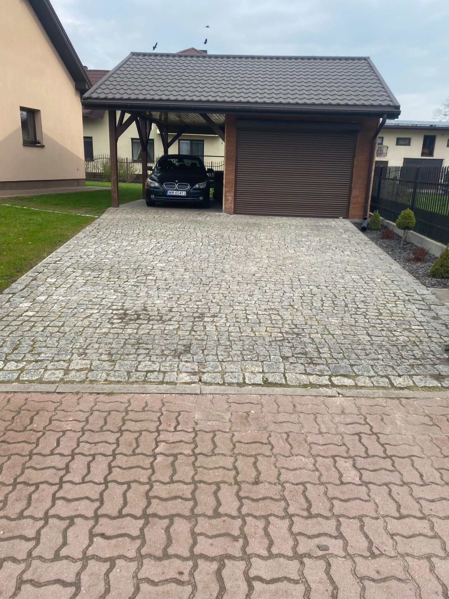 Podjazd z kostki granitowej prowadzący do garażu z samochodem. Widoczne obrzeża z kostki betonowej.