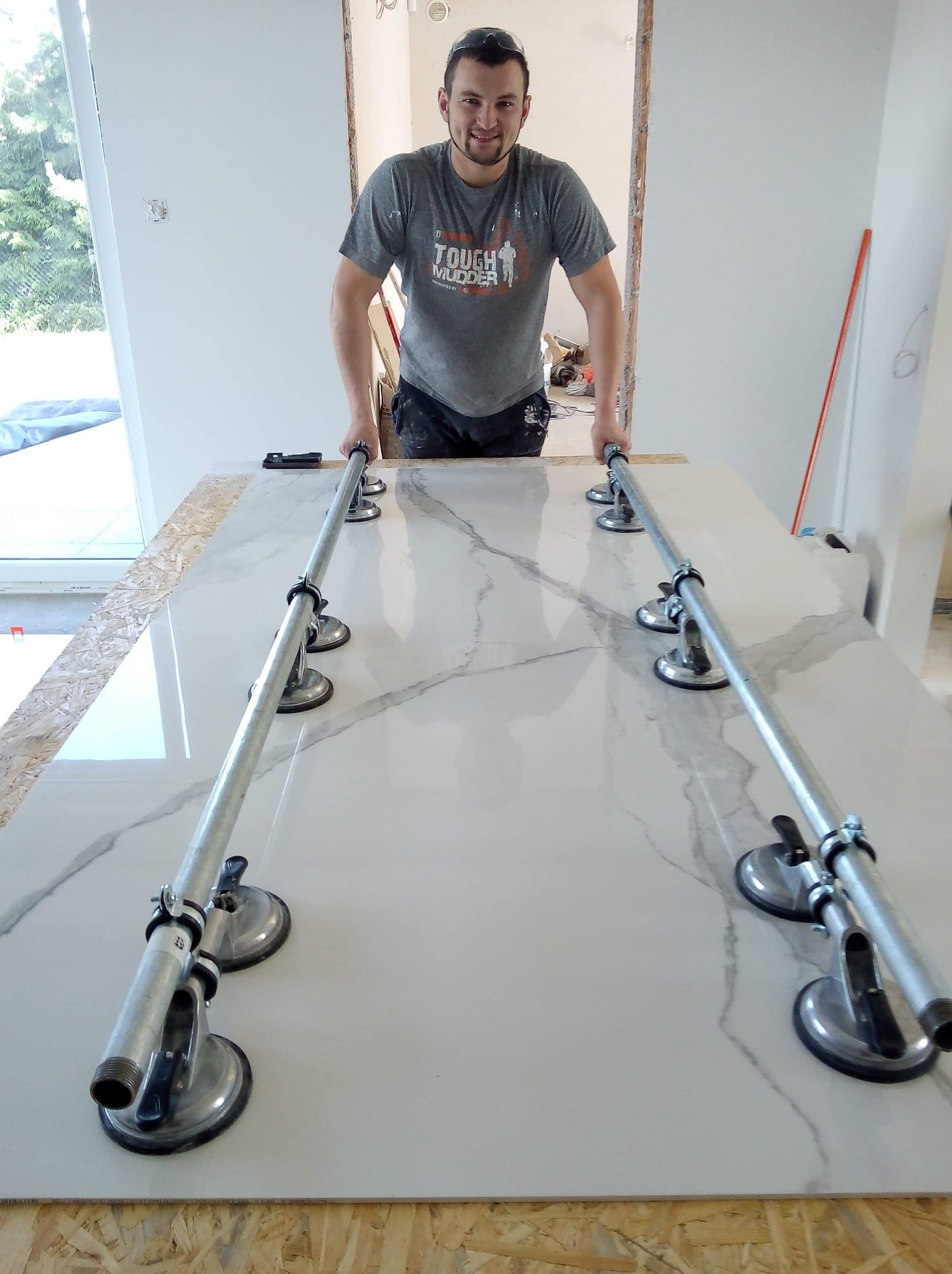 Glazurnik uśmiechnięty, przenosi dużą płytę gresową z marmurowym wzorem za pomocą specjalistycznych przyssawek na metalowych rurkach.