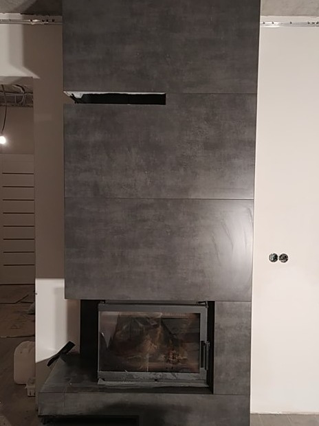 Wkład kominkowy obudowany płytami imitującymi beton, z widoczną wnęką wentylacyjną w górnej części obudowy, na tle surowego stanu ścian i instalacji.