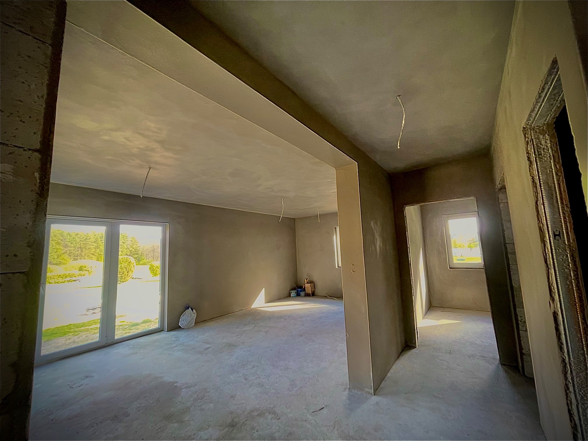 Surowe wnętrze w trakcie prac wykończeniowych: ściany pokryte świeżym tynkiem, widoczne przewody zwisające z sufitu, okna z widokiem na zieleń, podłoga w szarym betonie.