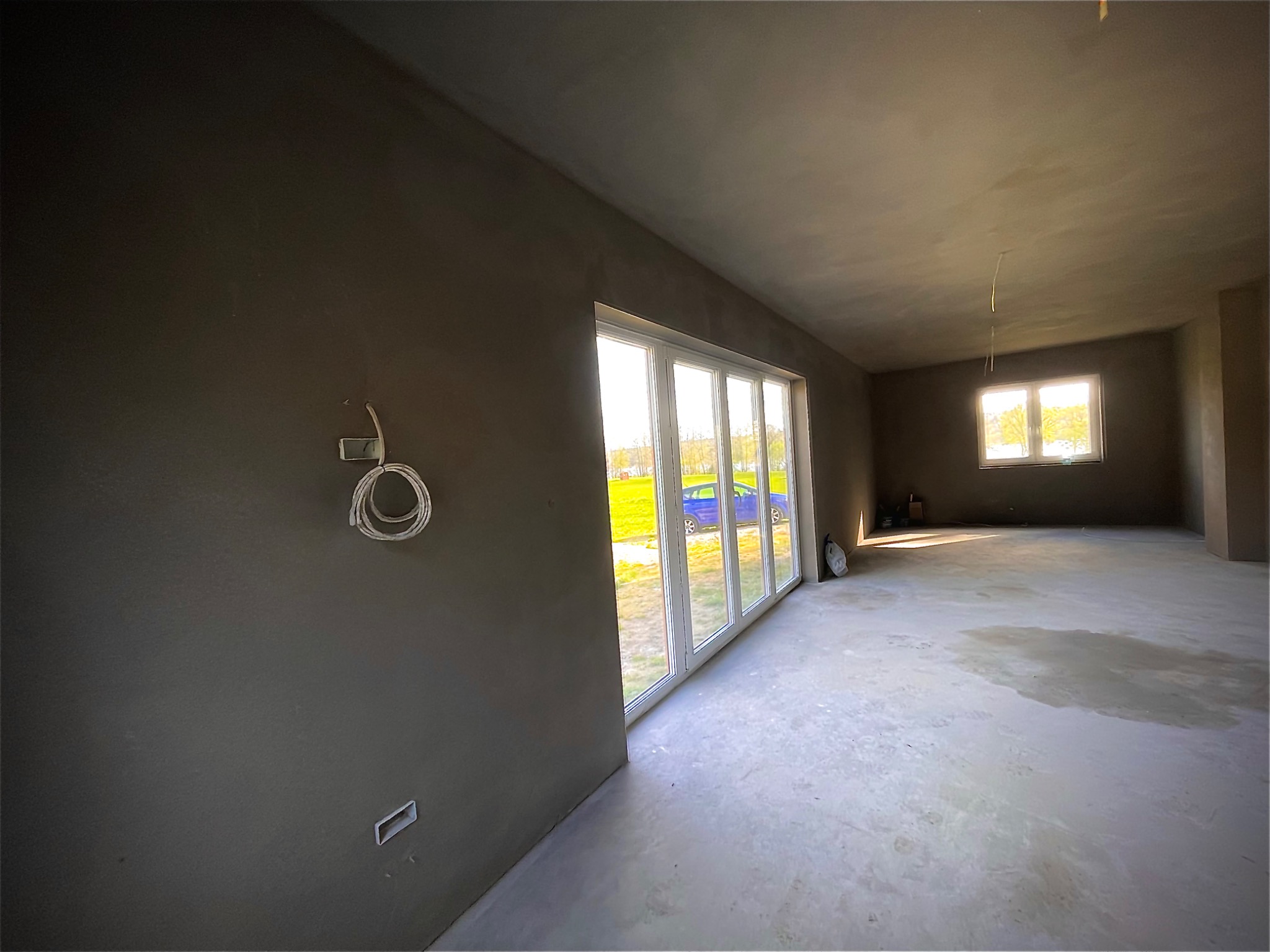 Surowe wnętrze w trakcie prac wykończeniowych: ściany i sufit pokryte szarym tynkiem, duże okno przesuwne z widokiem na zewnątrz, betonowa podłoga z widocznymi plamami, wystające kable ze ściany.