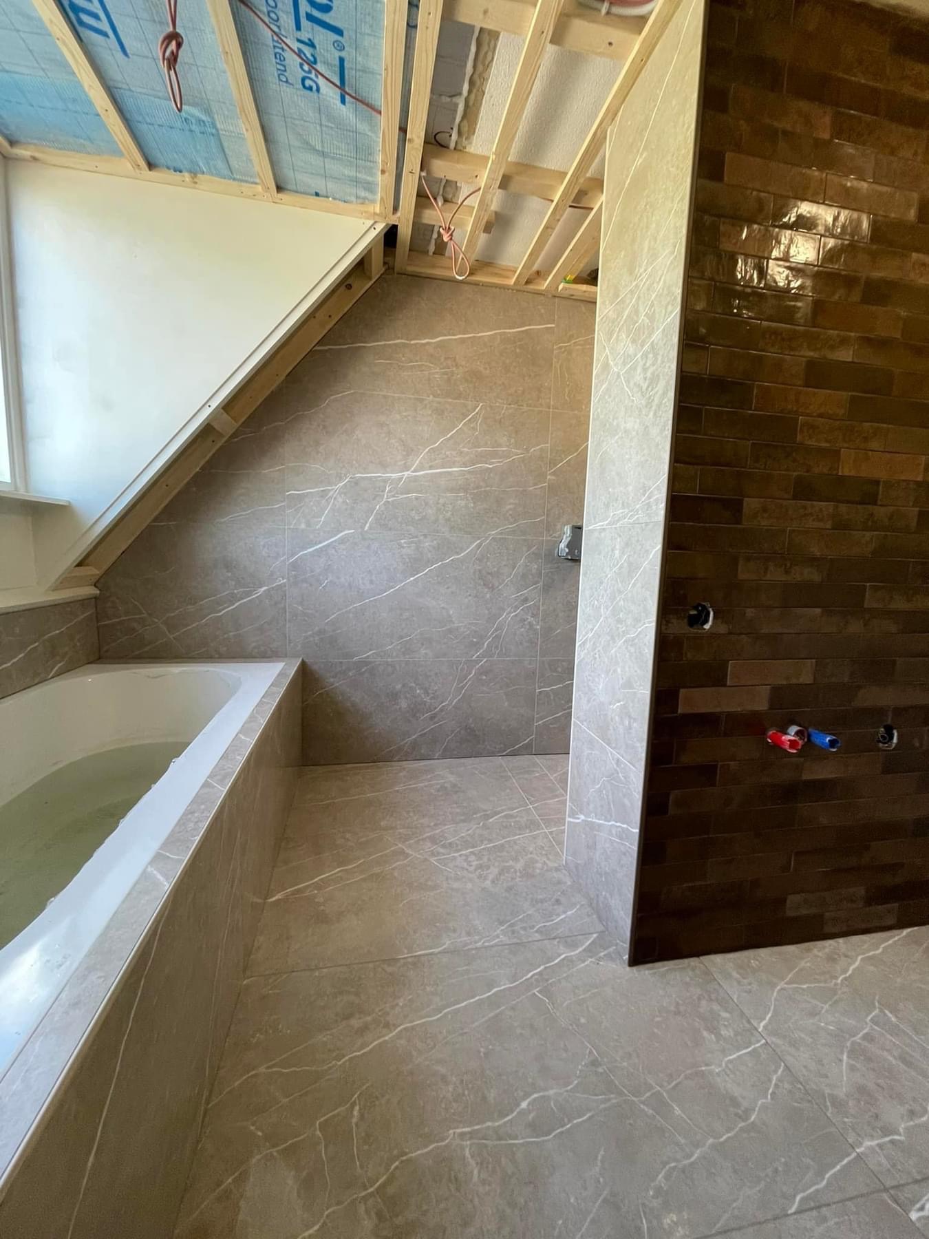 Wnętrze łazienki na poddaszu w trakcie wykańczania: ułożone płytki imitujące marmur na podłodze i ścianach, częściowo wykończona ściana z brązowej cegły, surowy sufit z drewnianą konstrukcją, wanna...