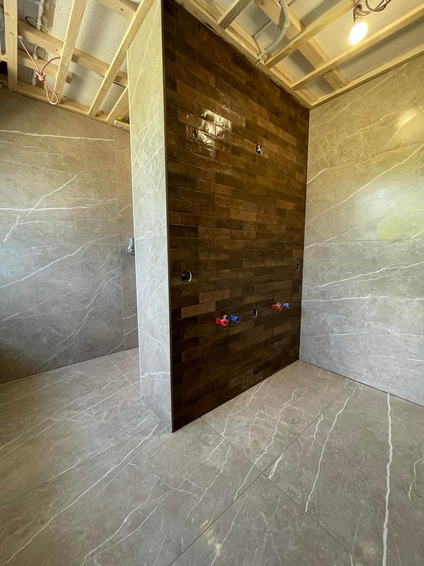 Łazienka w trakcie remontu: brązowe płytki na ścianie prysznica, szare płytki imitujące marmur na ścianach i podłodze, widoczne instalacje wodne i drewniana konstrukcja sufitu.