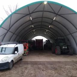 Duży namiot magazynowy z oświetleniem wewnętrznym, w środku zaparkowane maszyny rolnicze i biały samochód dostawczy z drabiną na dachu.