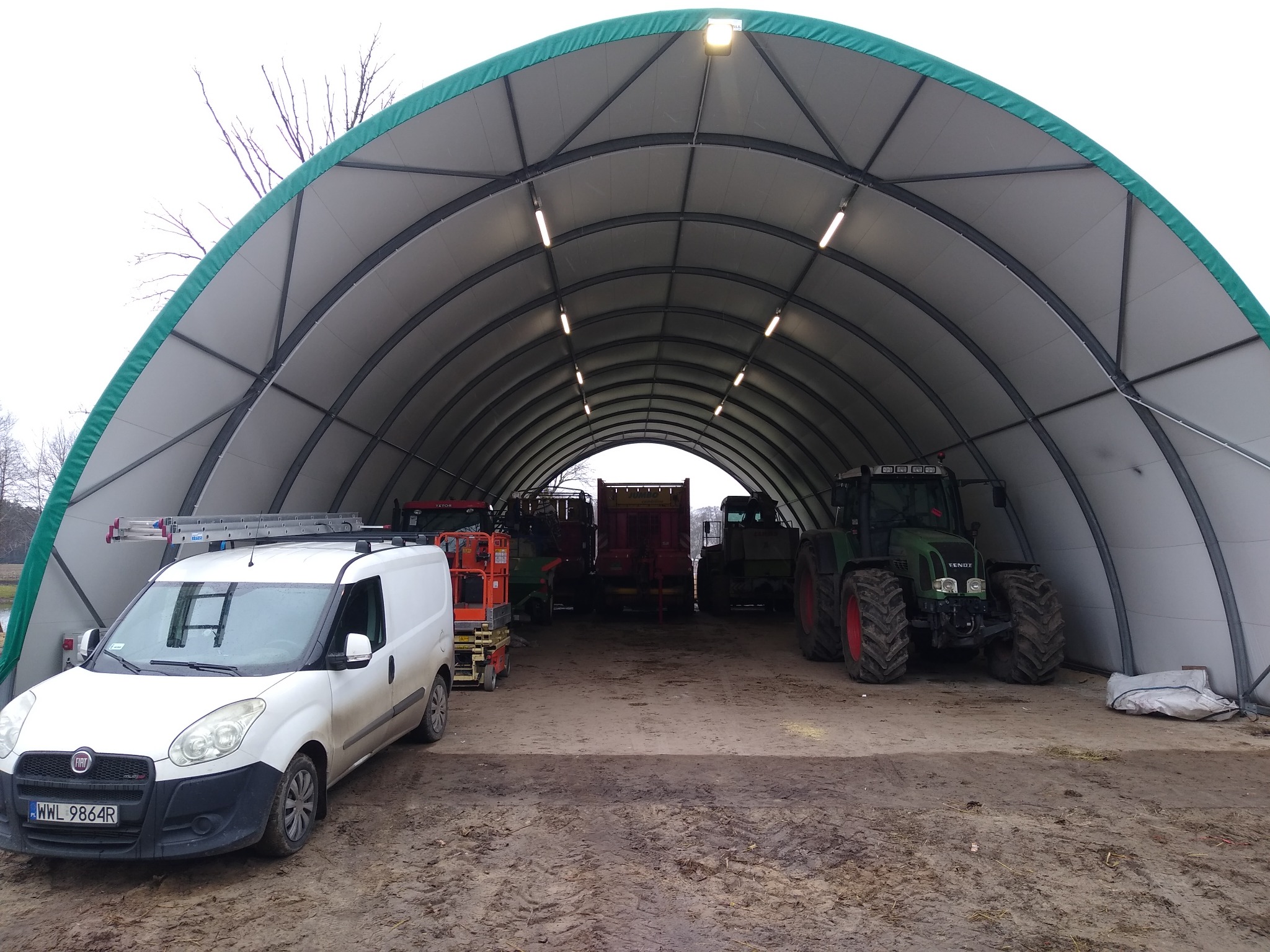 Duży namiot magazynowy z oświetleniem wewnętrznym, w środku zaparkowane maszyny rolnicze i biały samochód dostawczy z drabiną na dachu.