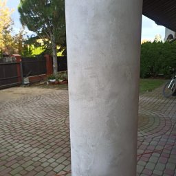 Quality House - Otynkowana kolumna na zewnątrz budynku z kostki brukowej w tle, widoczne drzewa i rower.