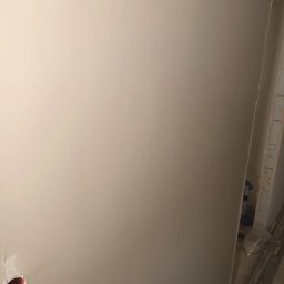 Quality House - Gładka, beżowa ściana po szpachlowaniu, widoczny fragment puszki elektrycznej w dolnym rogu i zabezpieczone folią elementy wykończeniowe w rogu pomieszczenia.