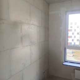 Quality House - Ściana z bloków betonu komórkowego z widocznymi zapiskami ołówkiem, częściowo pokryta wstępną warstwą gładzi, okno zabezpieczone folią malarską, widok na budynek mieszkalny za oknem.