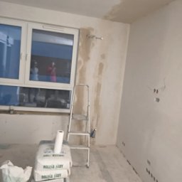 Quality House - Pokój w trakcie remontu, z widocznymi śladami po gładzi na ścianach, drabina oparta o ścianę, worki gładzi oraz rolka papieru na podłodze, okno z odbijającymi się postaciami.