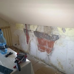 Quality House - Ściana w trakcie przygotowania do prac wykończeniowych, z widocznymi fragmentami cegieł i starej farby, zabezpieczona lampą roboczą i narzędziami na stole.