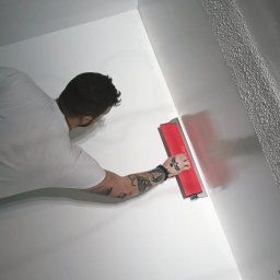 Quality House - Mężczyzna z tatuażami na ramieniu wygładza ścianę szpachlą z czerwoną rączką, widoczny fragment ściany z nałożoną, świeżą warstwą.