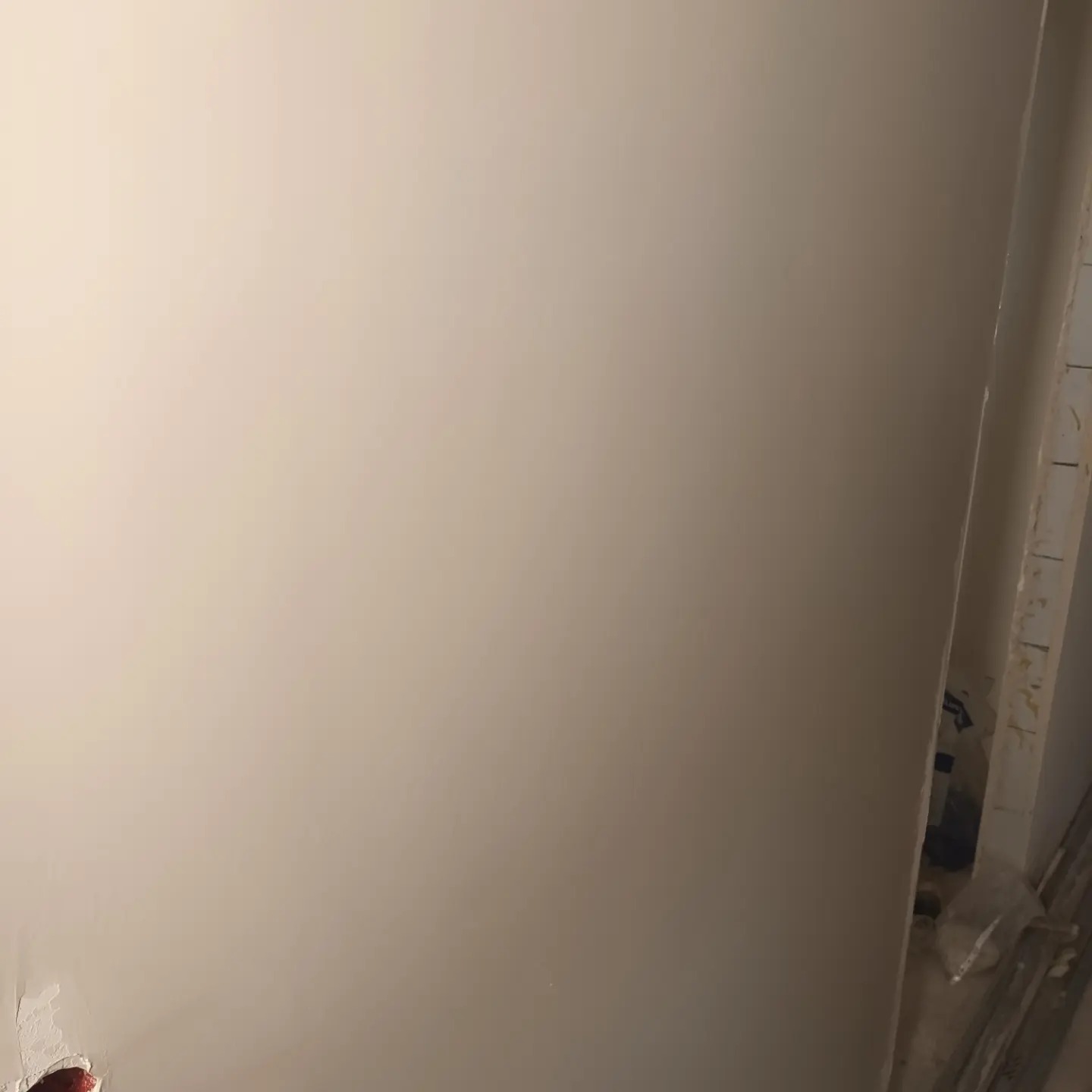 Gładka, beżowa ściana po szpachlowaniu, widoczny fragment puszki elektrycznej w dolnym rogu i zabezpieczone folią elementy wykończeniowe w rogu pomieszczenia.