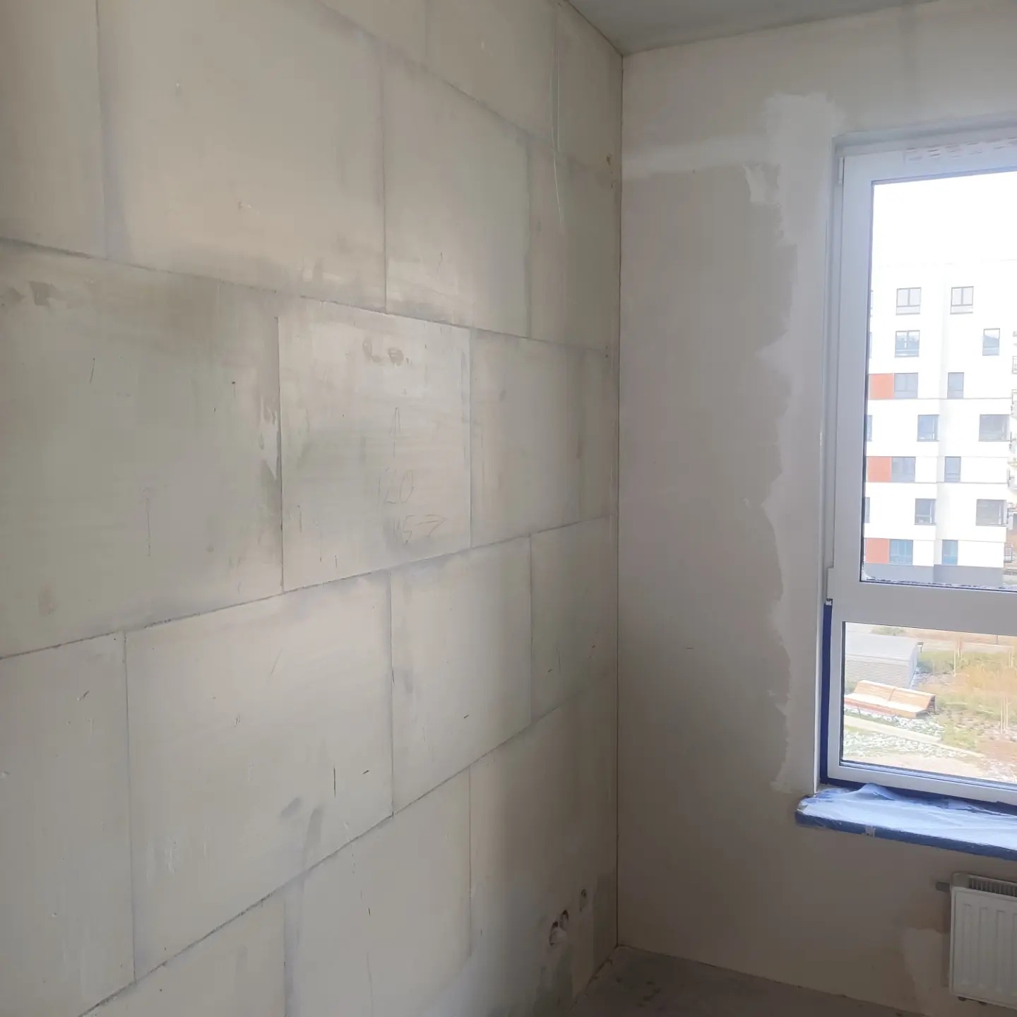 Ściana z bloków betonu komórkowego z widocznymi zapiskami ołówkiem, częściowo pokryta wstępną warstwą gładzi, okno zabezpieczone folią malarską, widok na budynek mieszkalny za oknem.