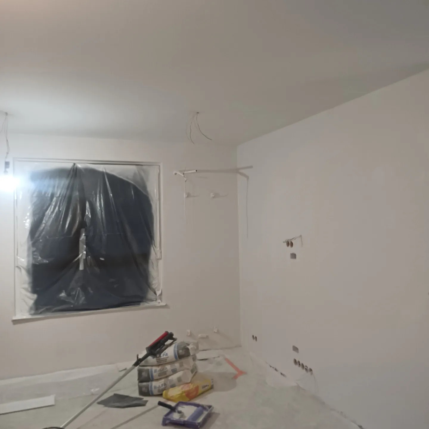 Pokój w trakcie malowania: okno zabezpieczone folią, worki z gładzią i narzędzia malarskie na podłodze, ściany przygotowane do malowania z widocznymi puszkami elektrycznymi.