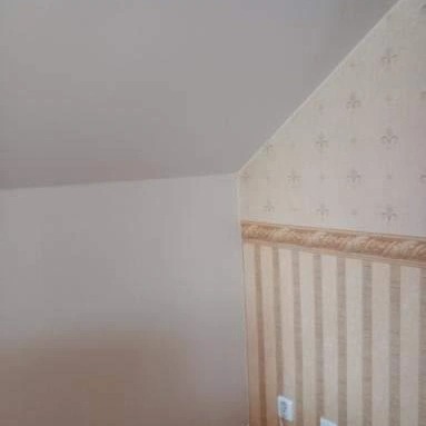 Ściana pod skosem po malowaniu na jasny kolor, sąsiadująca z fragmentem ściany pokrytej tapetą w beżowe pasy i wzory lilii, z widocznymi gniazdkami elektrycznymi przy podłodze.