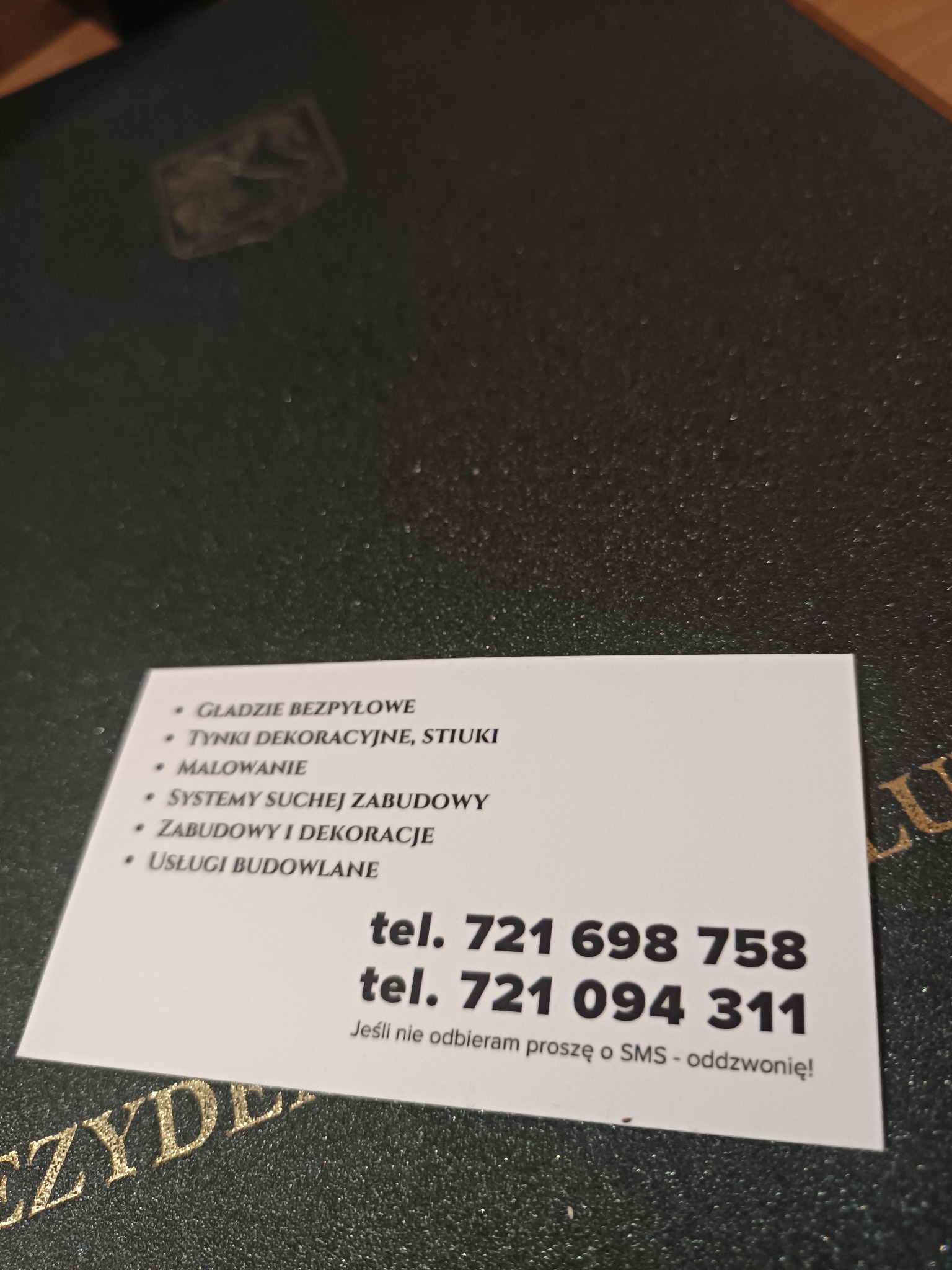 Wizytówka firmy budowlanej leżąca na ciemnym, teksturowanym tle. Wizytówka oferuje gładzie bezpyłowe, tynki dekoracyjne, malowanie, systemy suchej zabudowy, zabudowy i dekoracje oraz usługi...