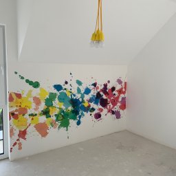 Rafbud - Białe wnętrze pokoju z kolorowymi plamami farby na ścianie, żółta lampa wisząca z trzema żarówkami i surowa podłoga.