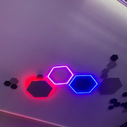 Rafbud - Sufit z dekoracyjnymi panelami LED w kształcie sześciokątów, świecących na czerwono, różowo i niebiesko, oraz dodatkowymi panelami w kolorze szarym i czarnym.