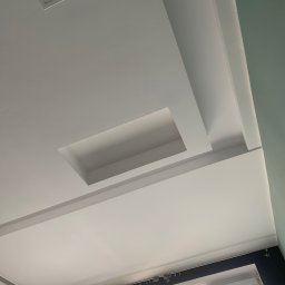 Rafbud - Biały, podwieszany sufit z wnękami na oświetlenie i dekoracyjnymi listwami przysufitowymi, widok z dołu na okno z żaluzjami i fragment ściany.