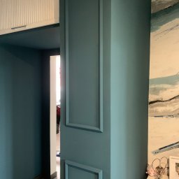 Rafbud - Ściana pomalowana na ciemny turkus z dekoracyjnymi, prostokątnymi listwami, widok na korytarz z szafką pod sufitem i fragment obrazu na ścianie