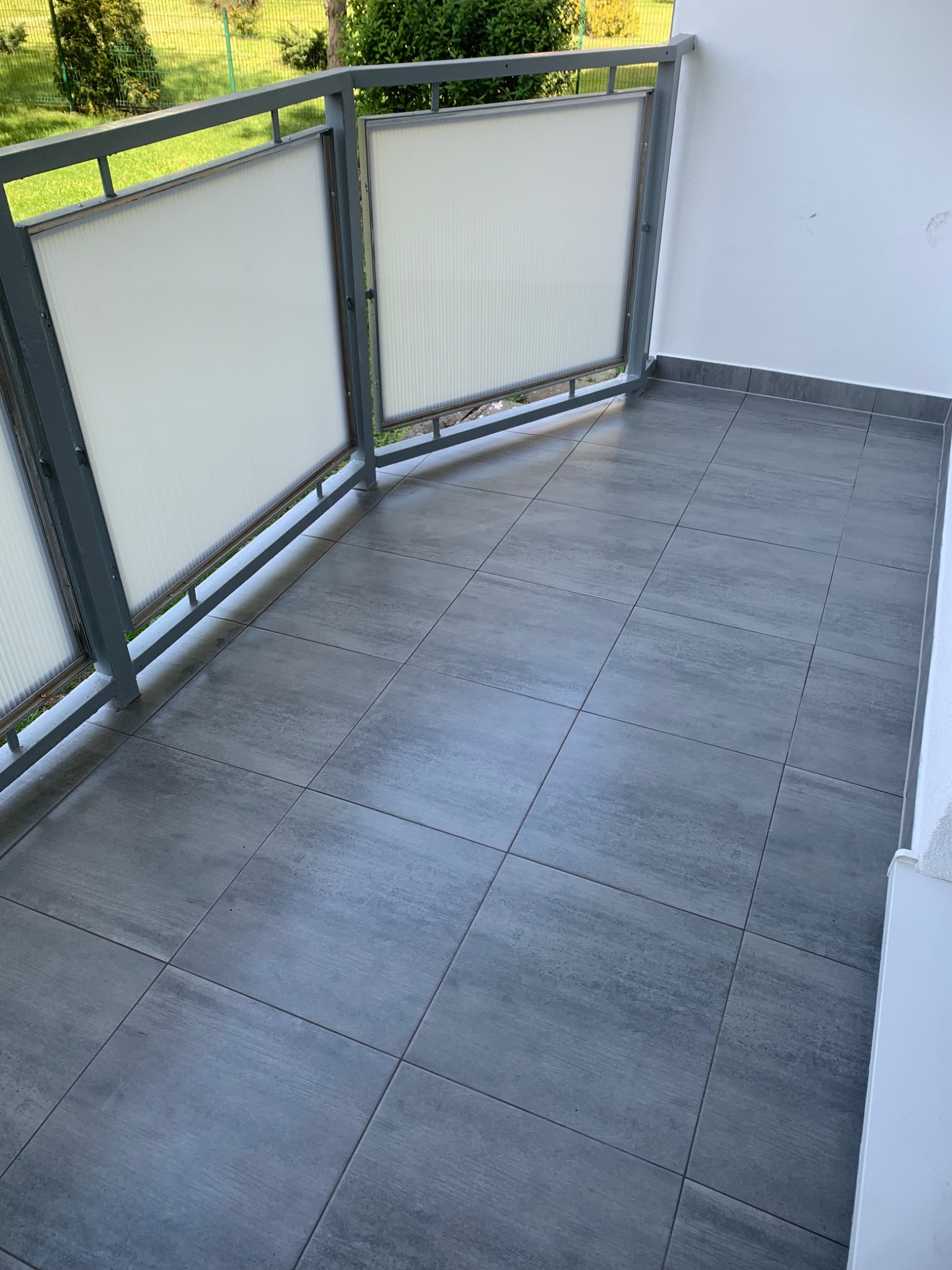 Wykończony balkon z szarymi płytkami podłogowymi, metalową balustradą z wypełnieniem z tworzywa sztucznego i widokiem na zieleń.