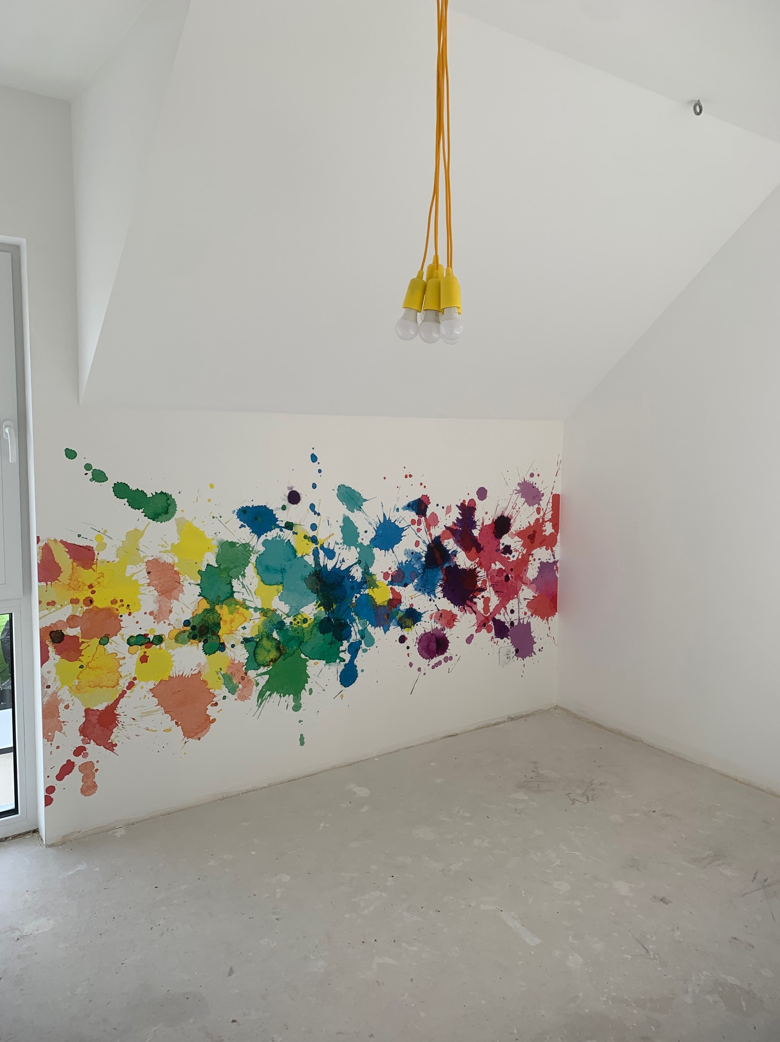 Białe wnętrze pokoju z kolorowymi plamami farby na ścianie, żółta lampa wisząca z trzema żarówkami i surowa podłoga.