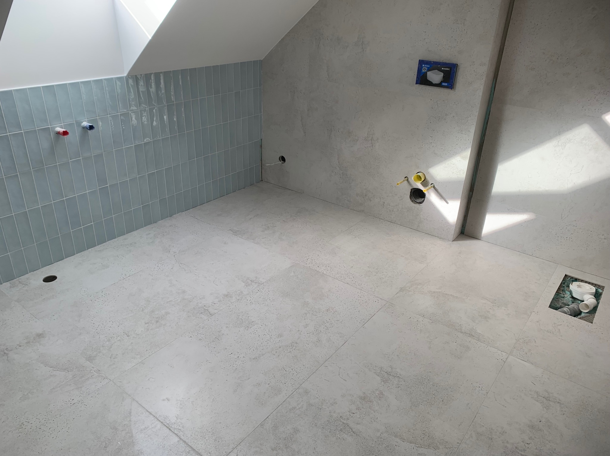 Wnętrze łazienki w trakcie prac wykończeniowych: jasne płytki podłogowe i na ścianie, widoczne instalacje hydrauliczne, sufit ze świetlikiem.