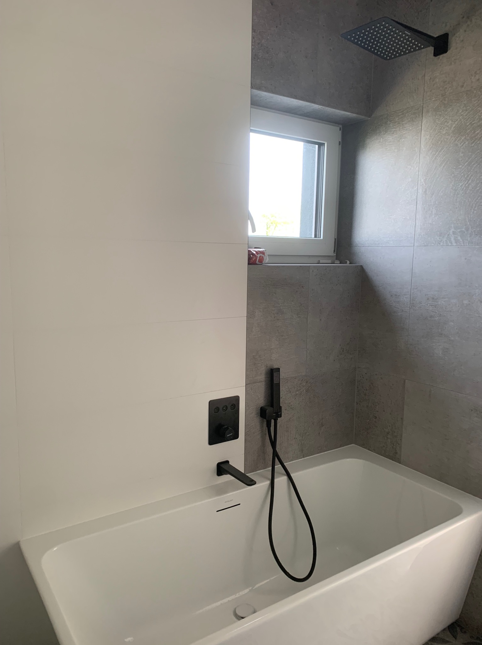 Nowoczesna łazienka z białą wanną, czarną armaturą i szarymi płytkami, widok na okno z dekoracyjnymi akcentami.