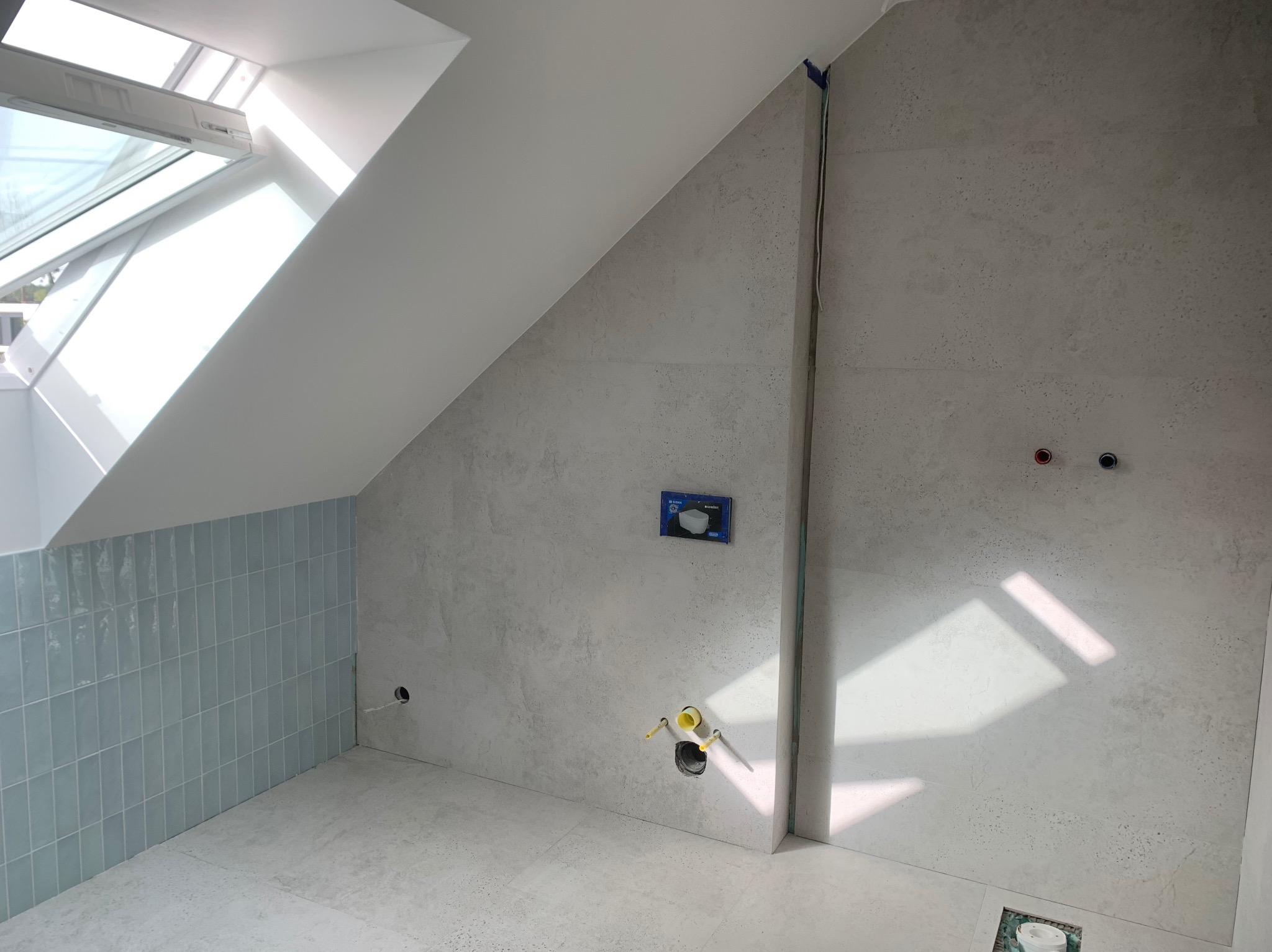 Wnętrze łazienki w trakcie remontu: ściany częściowo wyłożone błyszczącymi, pionowymi, błękitnymi płytkami, druga ściana z szarych płytek imitujących beton, widoczne wyprowadzone instalacje...