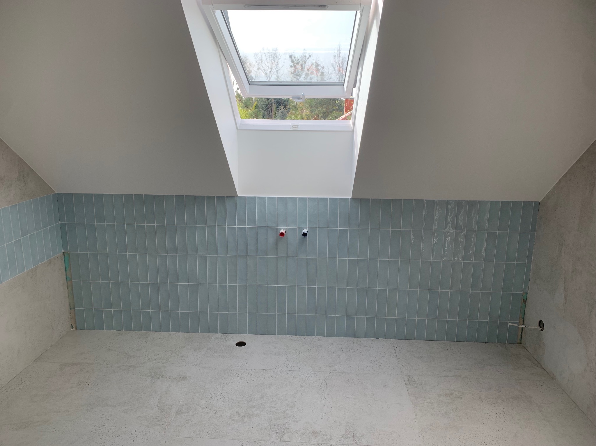 Świeżo położone, pionowe, błękitne płytki w łazience na poddaszu z oknem dachowym i przygotowanymi wyprowadzeniami na baterie oraz odpływem w podłodze.