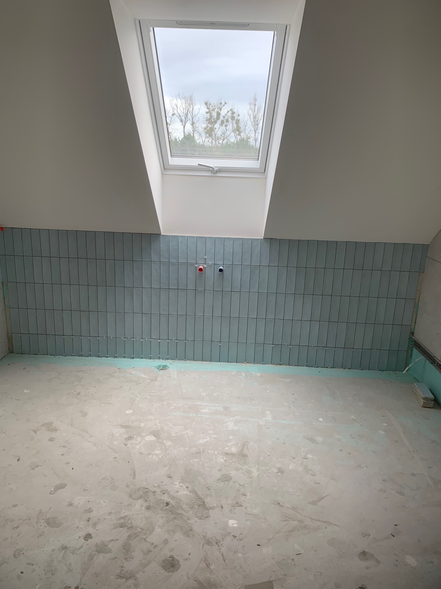 Łazienka na poddaszu w trakcie remontu: ułożone pionowo, błękitne płytki na ścianie, widoczne przyłącza wodne, okno dachowe i surowa podłoga.