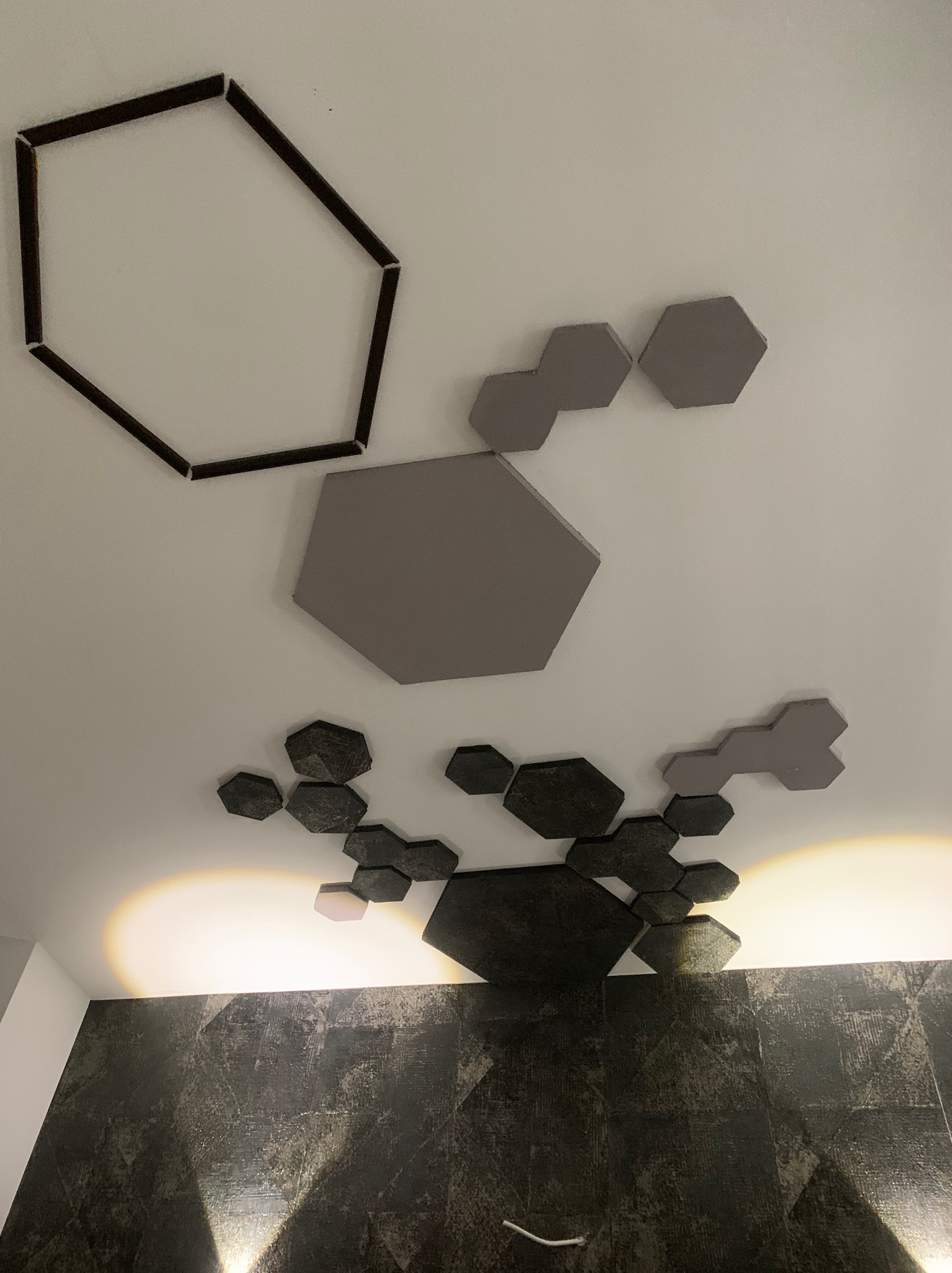 Geometryczne panele dekoracyjne w kształcie heksagonów w odcieniach szarości i czerni, umieszczone na suficie nad ciemną, teksturowaną ścianą.