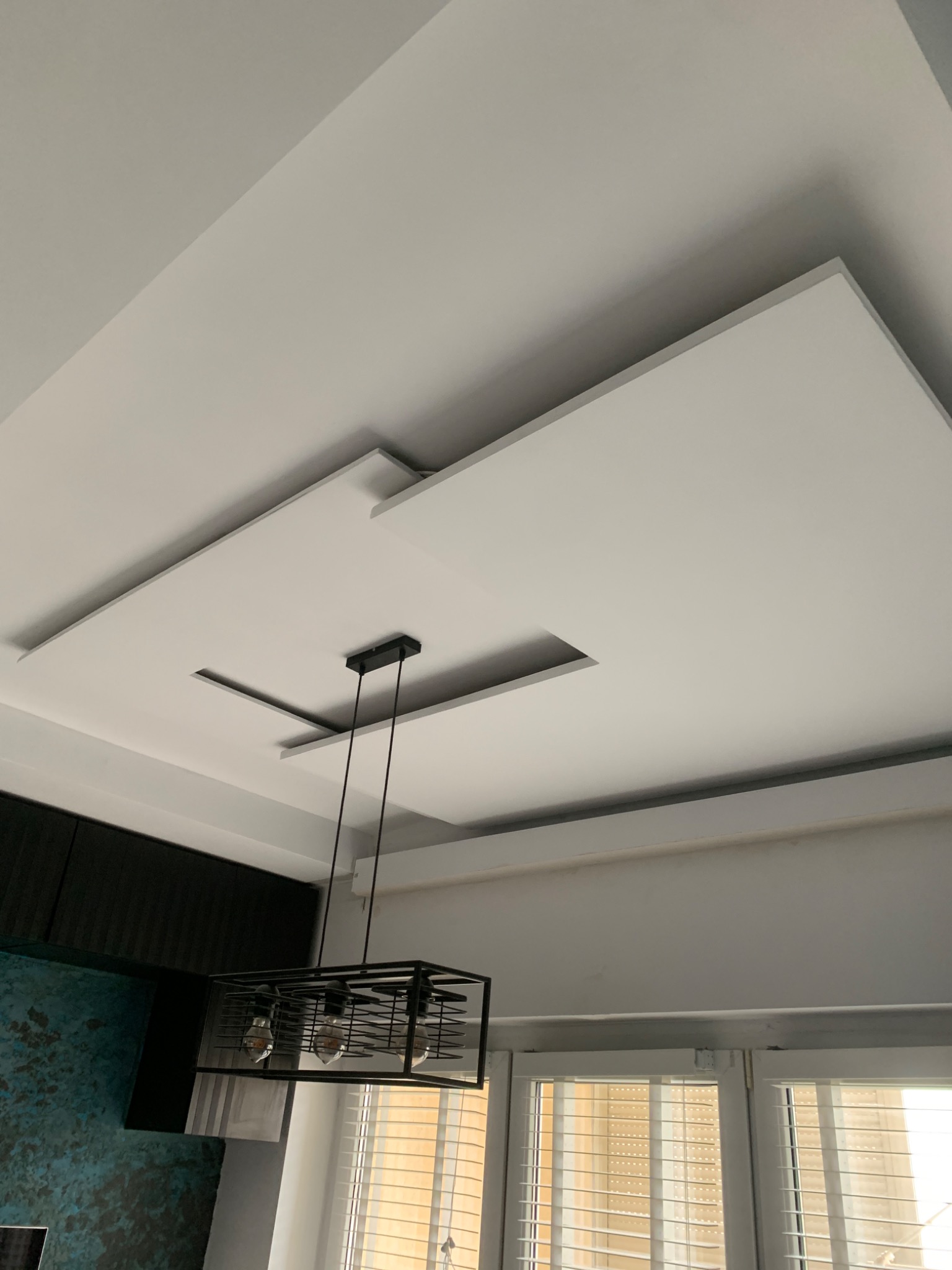 Wielopoziomowy sufit podwieszany z płyt kartonowo-gipsowych z wbudowanym oświetleniem i wiszącą lampą w metalowej klatce.