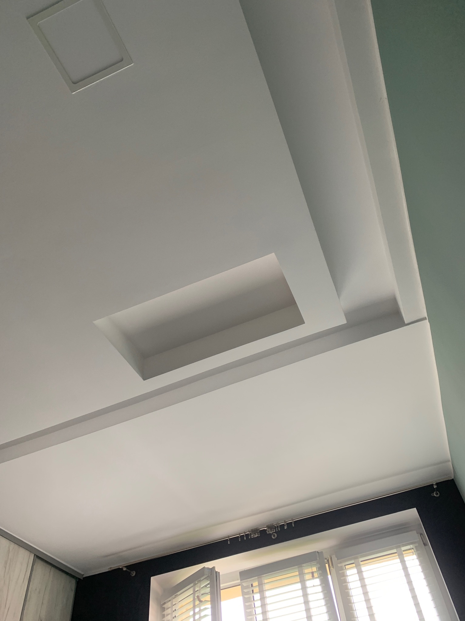 Biały, podwieszany sufit z wnękami na oświetlenie i dekoracyjnymi listwami przysufitowymi, widok z dołu na okno z żaluzjami i fragment ściany.
