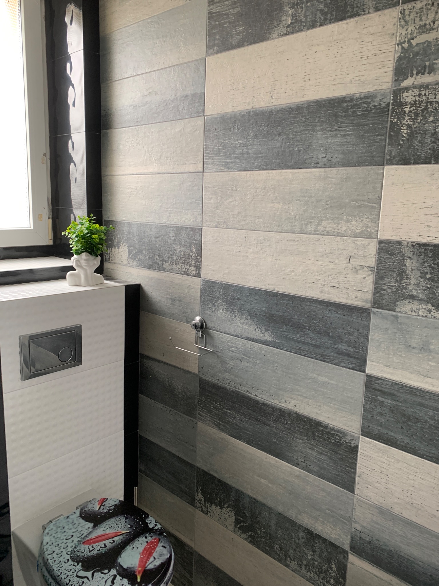 Wykończona łazienka z płytkami imitującymi drewno w odcieniach szarości i bieli, dekoracyjna roślina w białej doniczce w kształcie głowy, oraz deska sedesowa z motywem kamieni i kropli wody.
