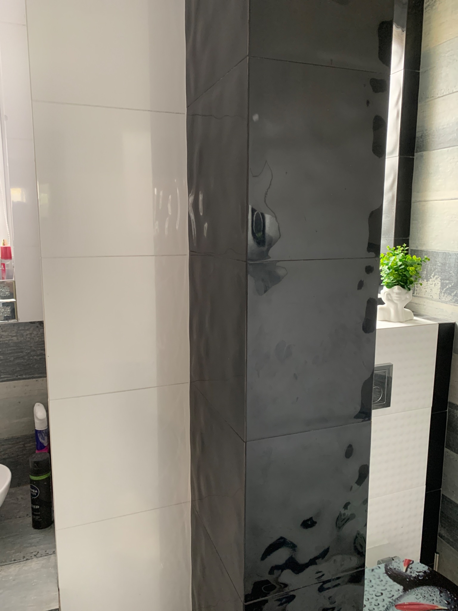 Fragment łazienki z czarno-białymi, błyszczącymi płytkami ceramicznymi ułożonymi w pionie, widoczna toaleta i dekoracyjna roślina w białej doniczce w kształcie głowy.