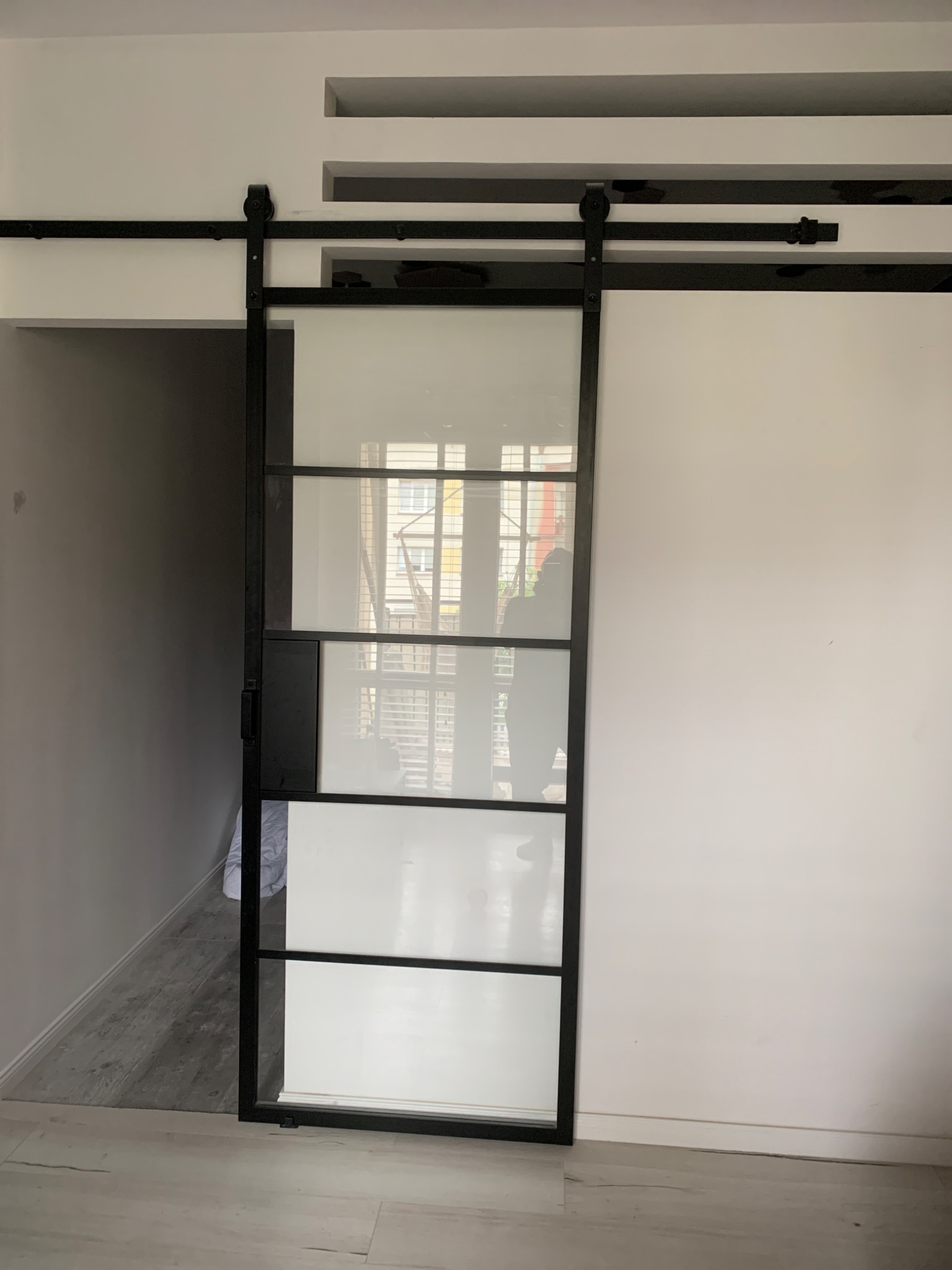 Nowoczesne drzwi przesuwne z czarną ramą i szklanymi panelami, zawieszone na szynie, oddzielające pomieszczenia w jasnym wnętrzu z dekoracyjnymi wnękami w suficie.