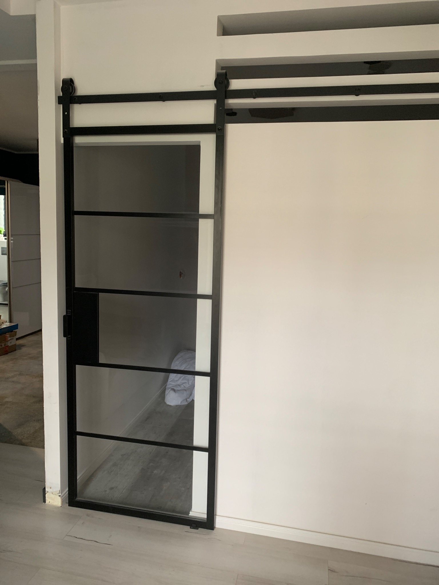 Przesuwne szklane drzwi w czarnej ramie z widocznym systemem przesuwnym na tle białej ściany. Widoczna podłoga z paneli w odcieniach szarości i fragment wnętrza pomieszczenia obok.