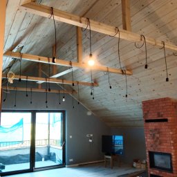 Jakub Ogar - Wnętrze poddasza z drewnianym stropem i widocznymi belkami konstrukcyjnymi, na których zawieszone są przewody z oprawkami żarówek, jedna żarówka świeci, przygotowanie do montażu oświetlenia, w tle...